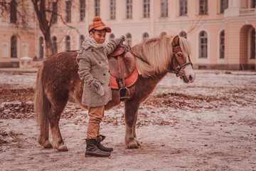 a boy stands next to a pony holding the saddle, the boy and pony look at the camera