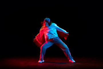 Young woman in casual clothes dancing hip hop isolated over black background in neon light with mixed lights effect. Concept of contemporary dance, street style, youth, hobby, action, lifestyle