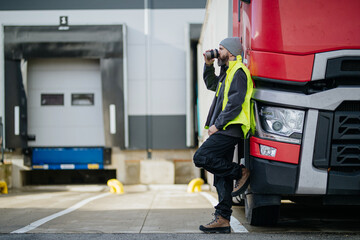 Truck driver leaning against red truck and drinking coffee, waiting for warehouse workers.