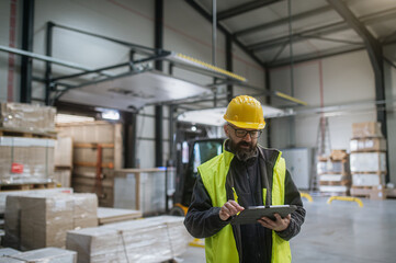 Warehouse receiver standing by delivered cargo, holding tablet, looking at cargo details, checking...