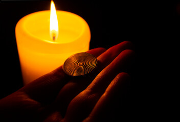 A woman's hand with a round golden disk on a burning candle. Dark dark background. Esoteric...