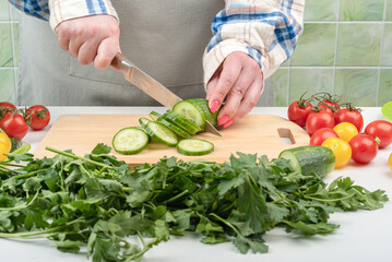 A young woman cuts a cucumber into slices on a kitchen wooden cutting board.
