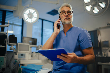 Portrait of confident ER doctor standing in hospital emergency room and phone calling. Handsome doctor in scrubs holding clipboard, standing in modern private clinic. - obrazy, fototapety, plakaty