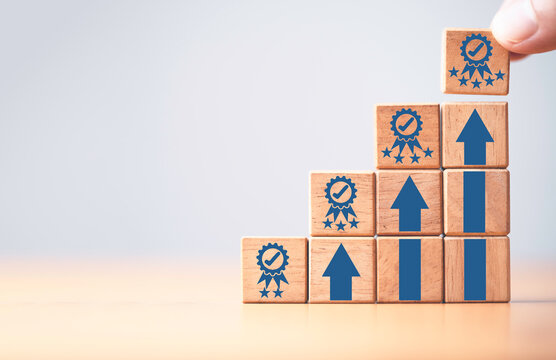 Hand holding the best quality guarantee symbol on the top of wooden stacking with up arrow for continuous quality improvement and customer satisfaction concept.