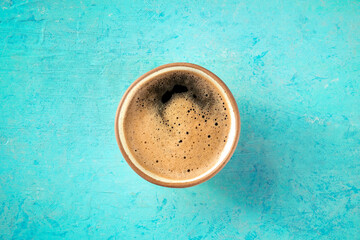 A cup of black coffee with froth, overhead flat lay shot on a turquoise background
