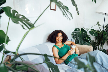 Beautiful laughing african american in casual clothes sits on sofa with cup of latte in her hands.