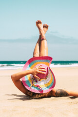 Woman with colorful hat enjoy the beach laying on the sand in summer holiday vacation travel lifestyle - young female caucasian people take a sunbath alone with blue ocean in background - obrazy, fototapety, plakaty
