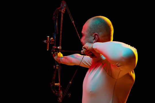 Back view image of man, archery at hele aiming with archery bow on target against black studio background in neon light. Concept of professional sport and hobby, competition, action, game
