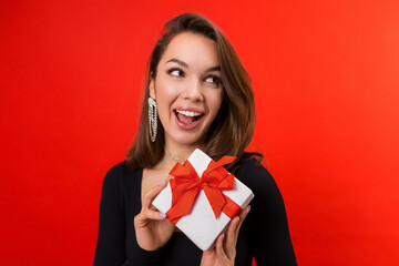 Fototapeta premium Valentine's day. A young woman gives a gift in a white box. Studio portrait on a red background