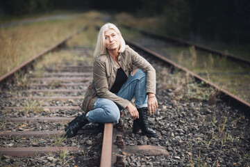 Naklejka na ściany i meble An older woman in her fifties with blonde hair, dressed in a brown jacket and jeans, sits with her legs bent on a railway track.