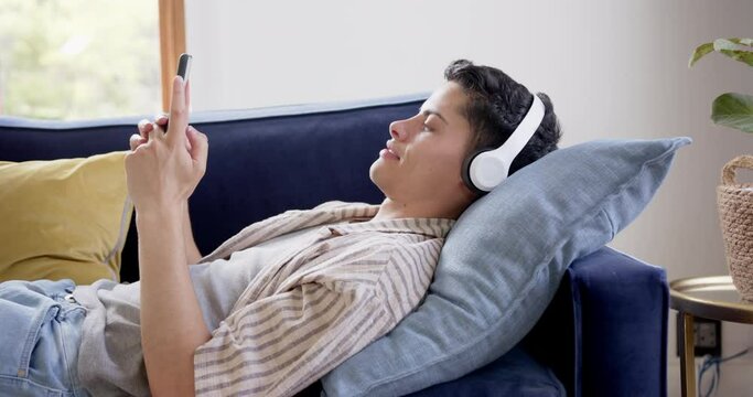 Happy biracial man in headphones lying on couch using smartphone, slow motion