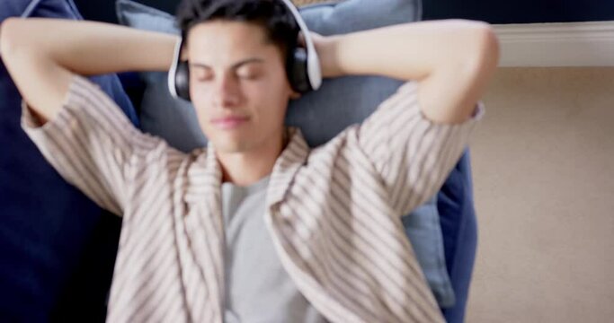 Happy biracial man wearing headphones lying on couch relaxing and smiling, slow motion
