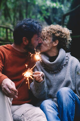 Romantic adult couple kissing in love holding fire sparklers sitting in the nature park enjoy outdoor relationship and leisure activity together. Concept of happy people enjoying vacations