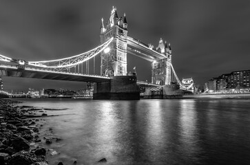 Tower Bridge ispanning over river Thames at evening twilight with bright illumination. Landmark, sight and tourist attraction in english metropole Lodnon from river bank. Black and white greyscale.