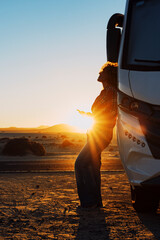 Happiness and freedom life with woman against camper van and golden sunset in background. People enjoying vanlife and summer holiday vacation with big camping car parked off road. Alone traveler