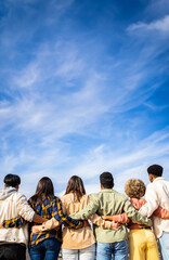 Obraz premium Vertical photo. Rear view of young group of happy multiracial friends hugging each other standing in a row looking blue sky