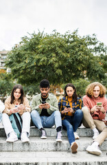 Fototapeta premium Vertical photo. Millennial group of diverse young people using mobile phone outside. Connected student friends on smartphone devices sitting in city street. Technology lifestyle concept