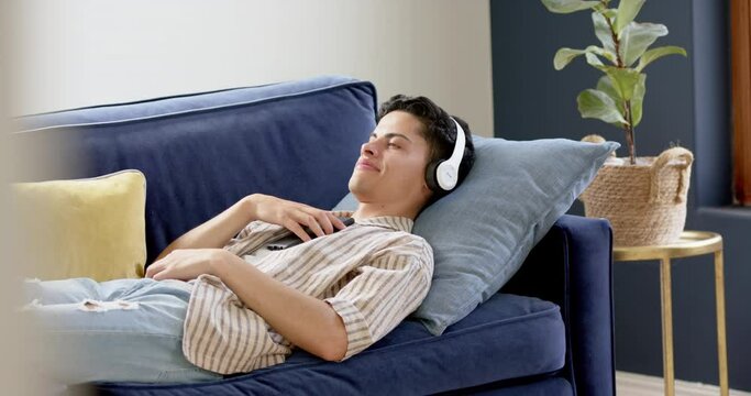 Happy biracial man in headphones lying on couch using smartphone, copy space, slow motion