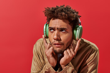 portrait of confused african american man in wireless headphones listening music on red background