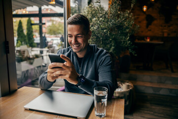 A happy man is sitting at coffee shop and smiling at his phone.
