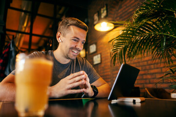 A happy man is sitting at coffee shop and having conference call while smiling at tablet.