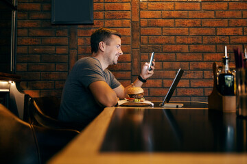 A trendy man is sitting at restaurant on a lunch break and smiling at his phone.