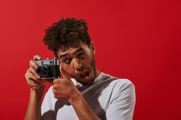 Fototapeta premium funny african american photographer looking at viewfinder while taking shot on red background
