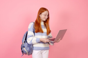 Smiling redhead teenage girl with bag and laptop wearing glasses on pink background in studio.