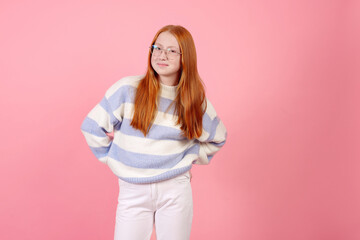 Fototapeta premium Curious redhead teenage girl wearing glasses and sweater on pink background in studio.