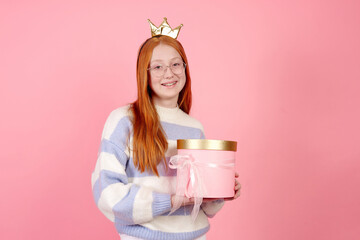 Smiling redhead teenage girl wearing glasses, crown and sweater holding present box on pink background in studio.