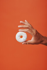 cropped view of person holding glazed vanilla donut with sprinkles on orange background, white icing