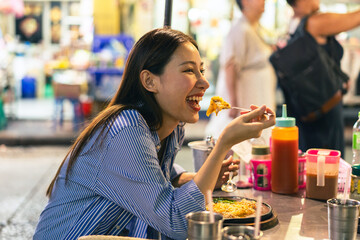 Young Asian woman traveler tourist eating Thai street food in China town night market in Bangkok in...
