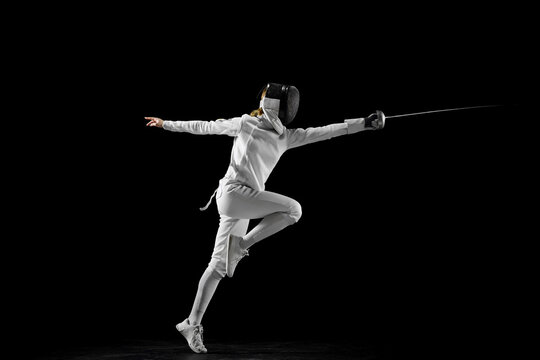 Confidence and strength. Female athlete in fencing gear showcasing her impeccable form, sword poised for action against a black studio background. Concept of active and healthy lifestyle, championship - Powered by Adobe