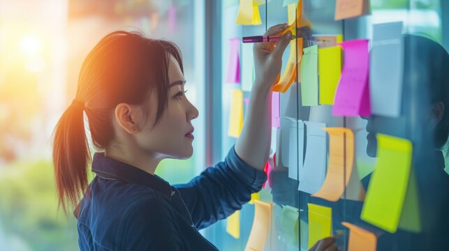 A woman standing in front of a wall full of colorful sticky notes. Young woman working in office and use post it notes to share idea. Communicating together in creative office. Brainstorming concept.