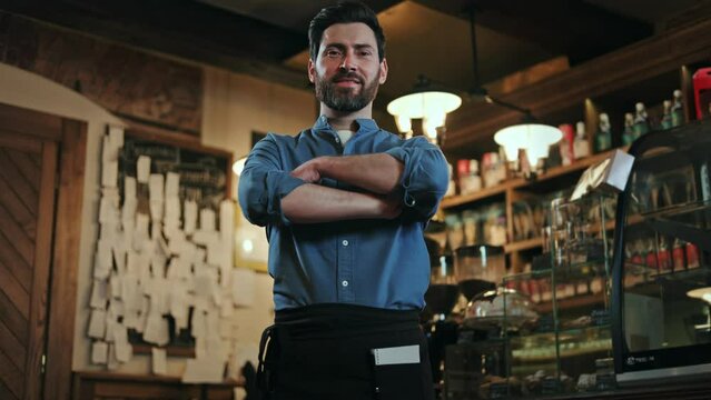 Low angle view of caucasian bearded man dressed in uniform looking confidently at camera and crossing arms. Qualified male waiter posing on background of bar counter full of alcohol bottles.