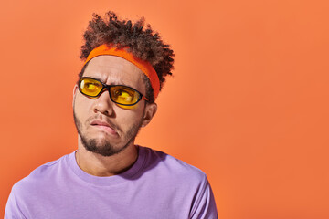 pensive african american man in sunglasses and headband looking away on orange background, grimace