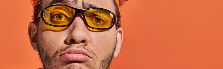 upset african american man in eyeglasses and headband grinning on orange background, grimace banner
