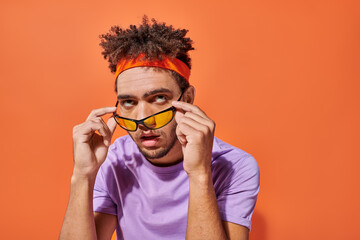 annoyed african american man in eyeglasses and headband rolling eyes on orange background, emotion