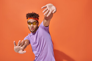 funny and expressive african american man in eyeglasses and headband gesturing on orange background