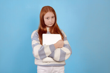 Fototapeta premium Confident redhead teenage girl holding laptop on blue background in studio.