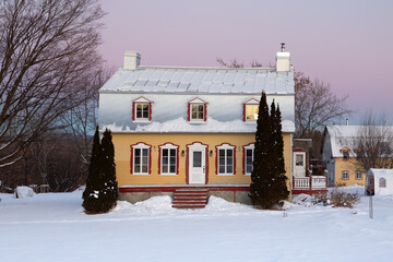 Sunrise winter view of beautiful patrimonial yellow clapboard house with silver metal Mansard roof and dormer windows, St. Pierre, Island of Orleans, Quebec