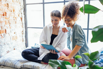 Two cute teenage girls are sitting on the windowsill and looking at the ipads screen.