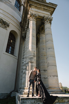 Newlyweds walk near Baroque Roman Catholic church in Pidhirtsi Ukraine. Bride and groom kissing on stairs near large columns of ancient temple at sunset. Couple hold hands nature autumn day.