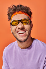 happy african american man in eyeglasses smiling with closed eyes on orange background, optimistic