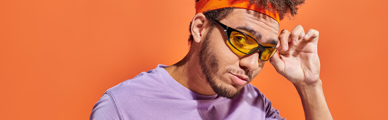 banner, young african american man adjusting sunglasses and looking at camera on orange background
