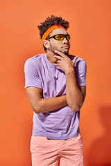 thoughtful young african american man in headband and sunglasses on orange background, funny face