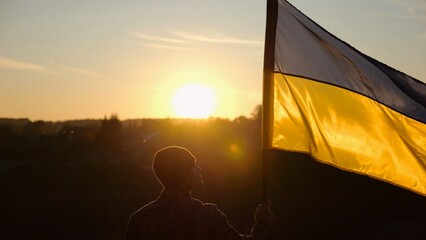Male soldier stands with raised flag of Ukraine against background of beautiful sunset. Male...