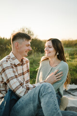 Happy young woman and man, walking spending time together in nature. Couple sitting on blanket, hugging in grass in field at sunset. Concept of family holiday outdoors. Female embrace male on picnic.