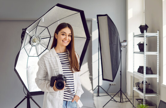 Young female photographer with digital camera posing in photo studio. Attractive smiling young woman holding professional dslr camera standing in studio with professional photographic equipment