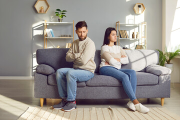 Family couple ignoring each other after a quarrel. Two people turn away and don't talk to each other. Angry young man and woman sitting back to back, with arms crossed on the sofa in the living room - obrazy, fototapety, plakaty